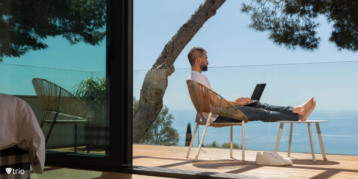 A man is working outdoors on a laptop, sitting on a bench with trees in the background. He is remotely accessing his work Mac, combining fresh air with productive work.