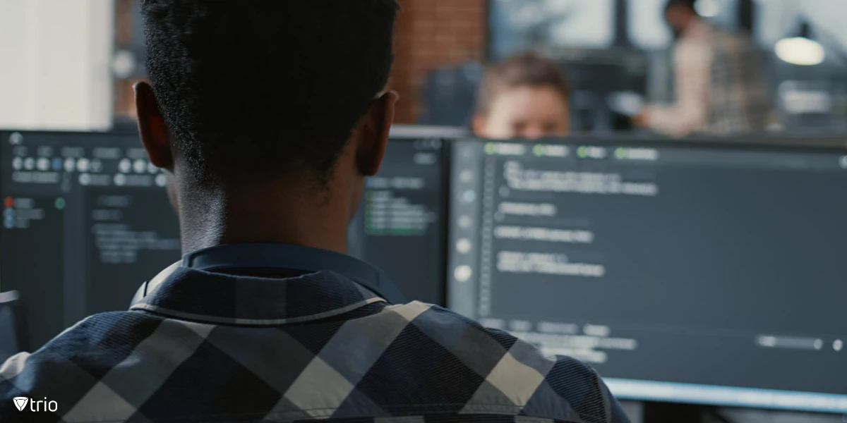 Portrait of programmer with laptop sitting at desk with multiple screens parsing code