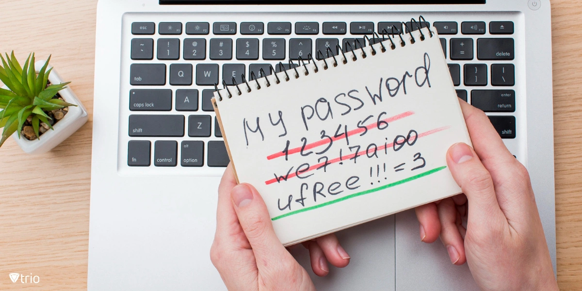 Hands holding a notebook displaying passwords next to a laptop.