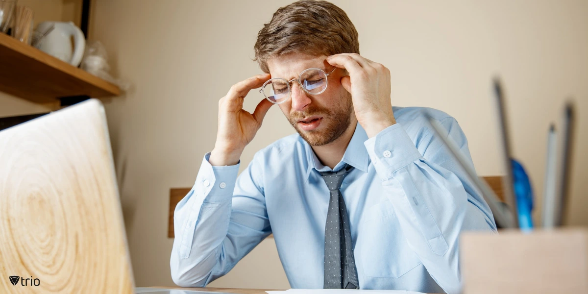 Frustrated office worker massaging his head while staring at his computer, overwhelmed by multiple MFA push notifications disrupting his workflow.