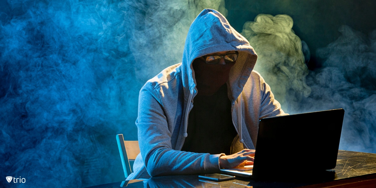 A hacker in a gray hoodie, with a mask and glasses, behind a laptop. The room is full of smoke