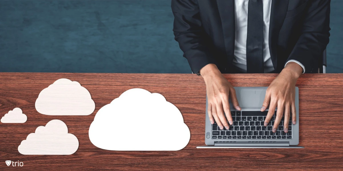 A person in business attire working on a laptop at a wooden desk. There are several white cloud shapes placed on the desk, symbolizing cloud computing or cloud storage