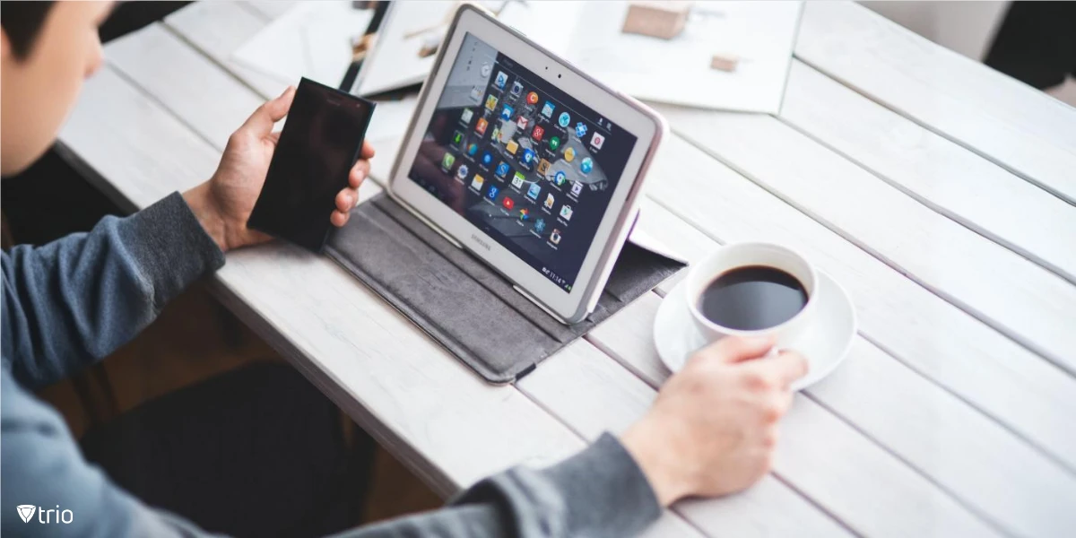 Man working on a desk with a tablet showing multiple apps and holding a mobile in one hand and a cup of coffee in the other