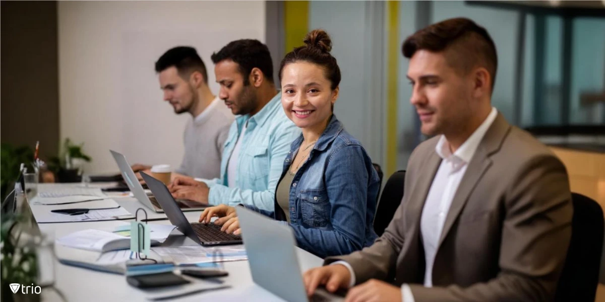 A group of individuals in a workspace, with a woman smiling at the camera while working on her laptop, surrounded by focused coworkers at a shared desk