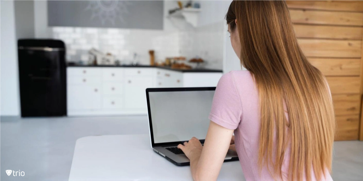 A person is sitting at a table in a kitchen, using a laptop