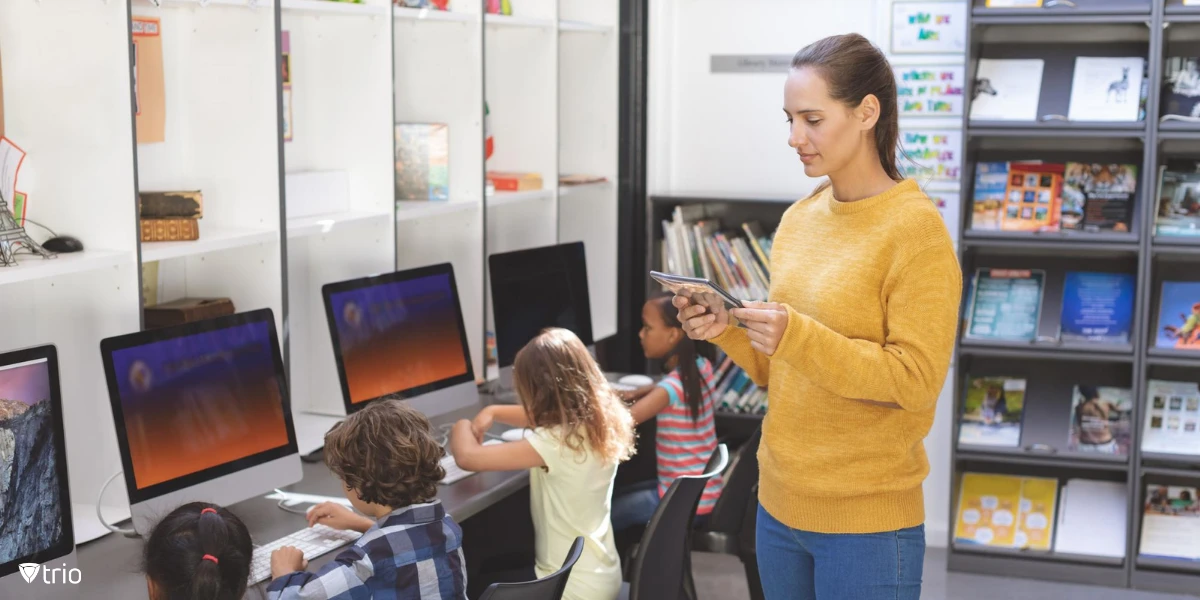 Ein Lehrer steht vor einer Reihe junger Schüler, die Desktop-Computer verwenden und ein Tablet in der Hand halten, um ihre Aktivitäten zu überwachen, in einer Schulbibliothek oder einem Klassenzimmer, mit Bücherregalen im Hintergrund