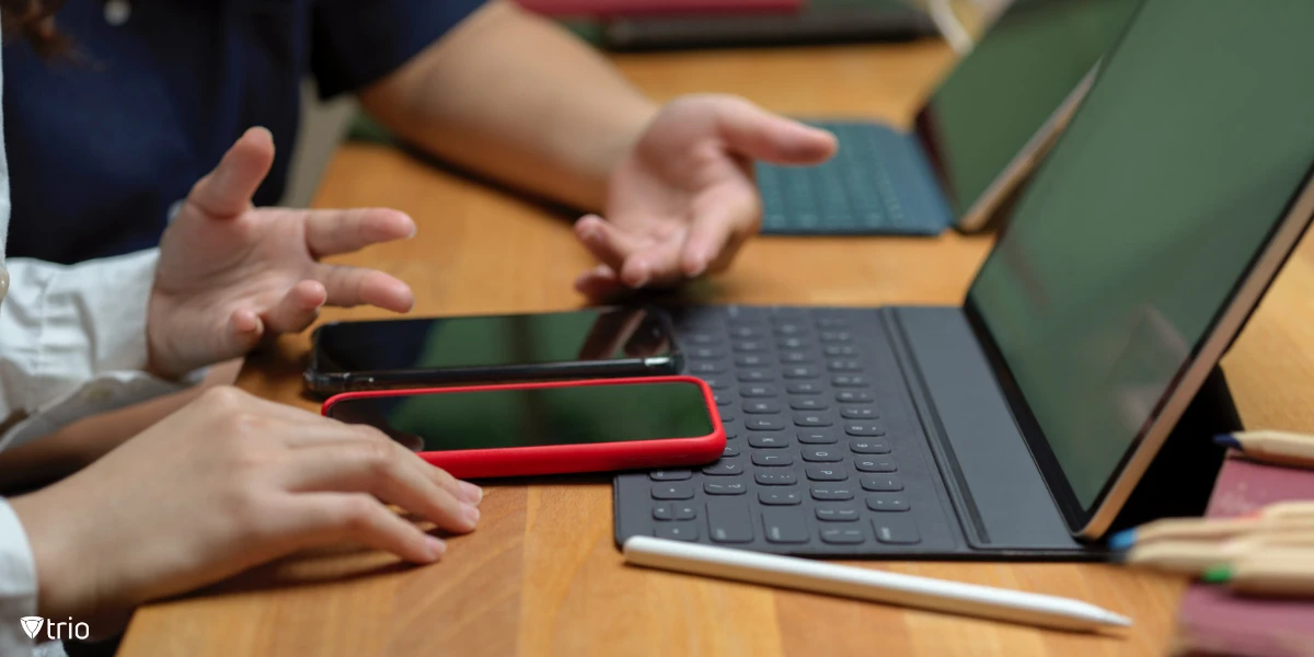 Die Hände von zwei Personen ruhen auf einem Holztisch, neben einem Tablet mit Tastaturaufsatz und mehreren Smartphones