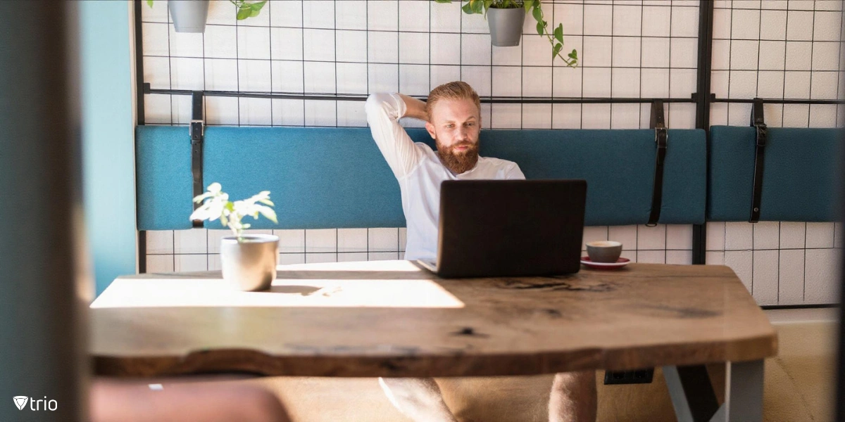 Ein bärtiger Mann mit hellem Haar, der an einem Holztisch sitzt, ein weißes Hemd trägt und an einem Laptop arbeitet, während er sich zurücklehnt und eine Hand hinter dem Kopf ruht.