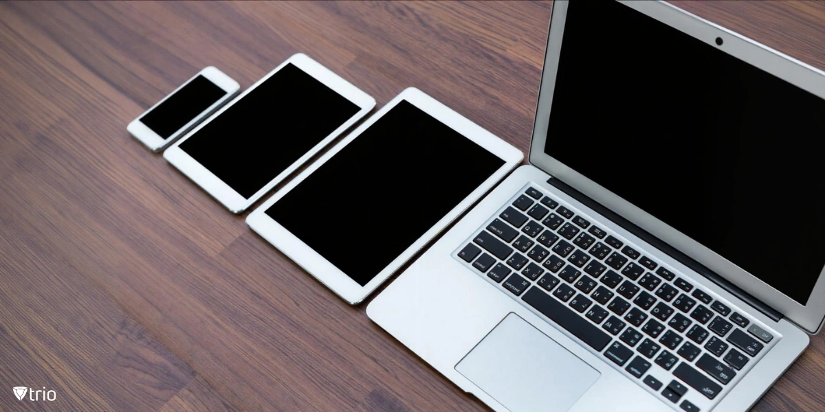 A collection of electronic devices arranged in a row on a wooden table. The devices include a smartphone, two tablets of different sizes, and a laptop