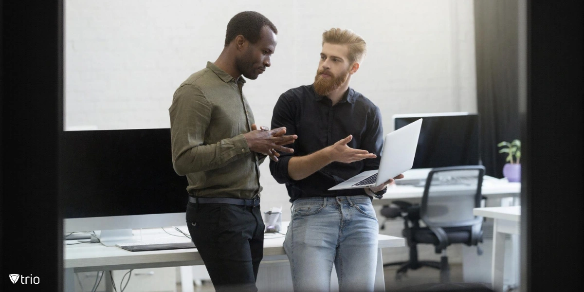 Two men in a modern office setting engaged in a discussion. One man is holding a laptop, while the other appears to be gesturing as they converse