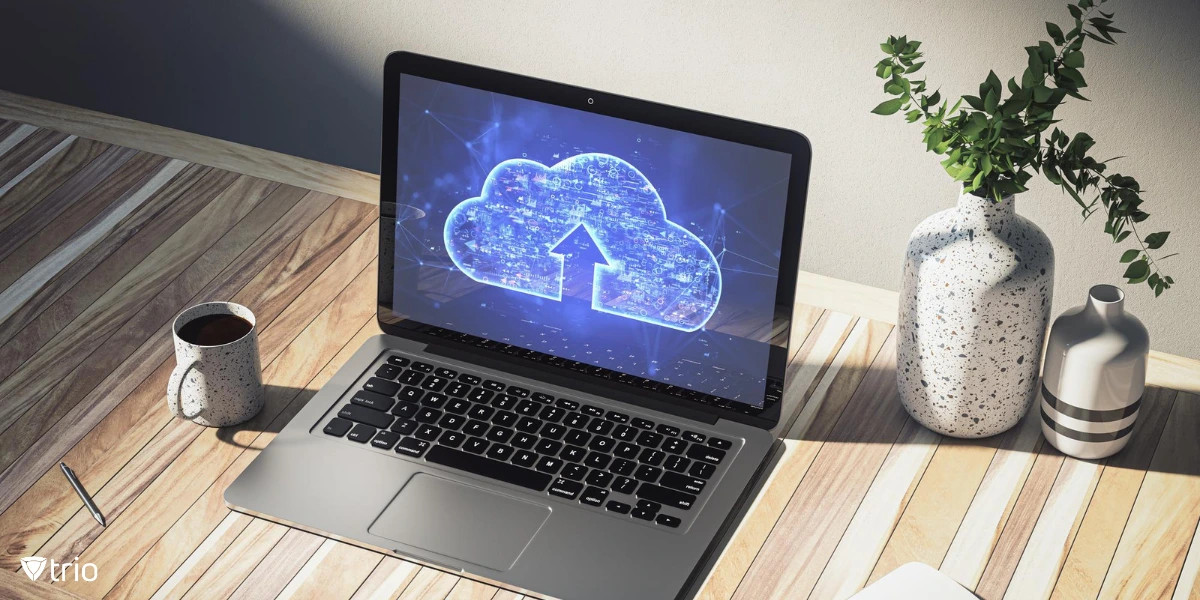 laptop sits on a wooden desk with a cloud upload symbol displayed on its screen, accompanied by a cup of coffee, a notebook, and decorative vases