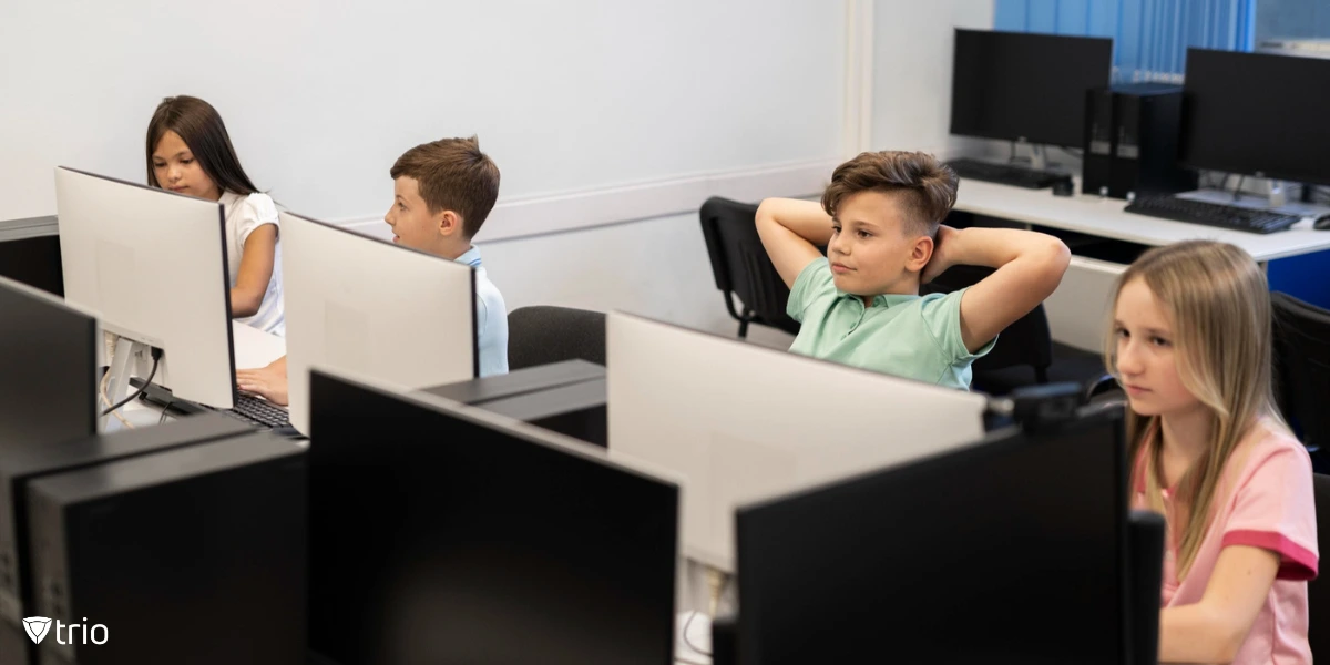 A group of young students is seated in a computer lab, working on desktop computers. One boy in a green shirt is leaning back with his hands behind his head, while others are focused on their screens. The room is well-lit, with multiple computer monitors visible